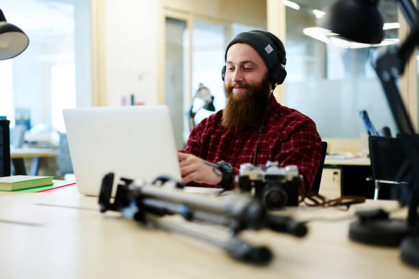 istock Positive male creating music using application on laptop computer and enjoying made composition via headphones.Bearded man in cool hat listening favorite playlist sitting in coworking 907948612