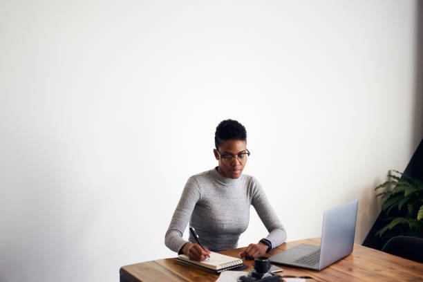 istock Concentrated Afro-American female designer creating sketch in notebook concentrated on ideas, professional businesswoman in eyewear planning projects making notes sitting on promotional background 907952162