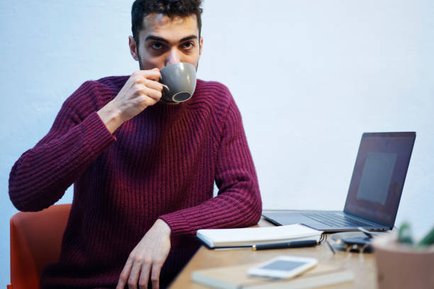 istock Handsome bearded hipster guy drinking tasty coffee in morning and looking away while working at modern laptop computer with sitting cozy cafe interior with copy space area 907954408