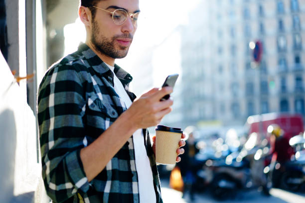 istock Serious handsome man in spectacles carefully watching interesting video on telephone connected to 4G internet while standing in sunshine urban setting.Traveler resting with modern device and coffee 907954410
