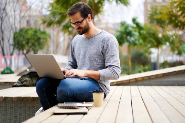 istock Positive bearded hipster guy in optical eyeglasses sitting in campus interior with coffee and typing text message on keyboard of modern laptop computer using high speed 4G internet connection 907961108