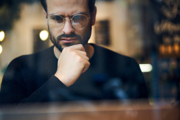istock Cropped close up image of pensive bearded entrepreneur in optical spectacles for better views dressed in black sweatshirt.Pondering businessman in trendy eyeglasses thinking on blurred background 907963392