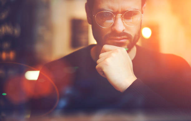 istock Cropped close up image of pensive bearded entrepreneur in optical spectacles for better views dressed in black sweatshirt.Pondering businessman in trendy eyeglasses thinking on blurred background 907963436