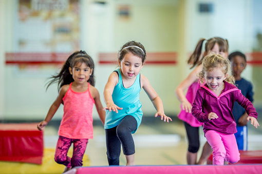 A group of preschool kids are indoors in their school gym. They are wearing casual clothes. Three girls are running and leaping onto a gymnastics mat.
