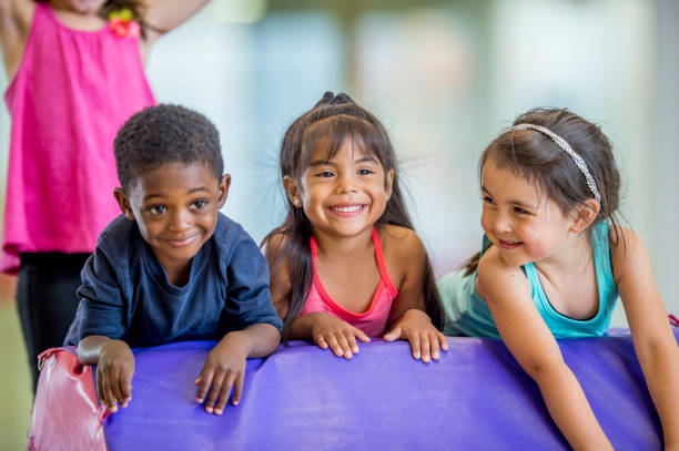 Kids In Gym Class Three preschool kids are indoors in their school. They are having fun during gym class. They are leaning on a gymnastics mat and smiling together. gymnastics stock pictures, royalty-free photos & images