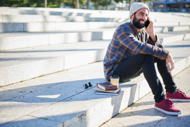 istock Smiling bearded guy having friendly talking on phone with friends enjoying weekend and spring weather sitting on urban setting background, cheerful male tourist using good connection in roaming 907970652