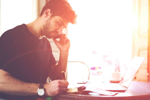 istock Male student communicating via telephone and writing down text information in notepad sitting at laptop computer in coffee shop interior.Bearded male having mobile  conversation via cellular 907970916