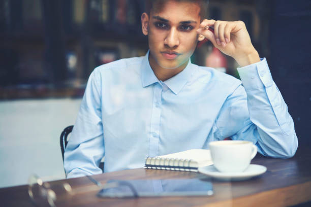 istock Cropped image of thoughtful young hindu author thinking on writing poems with interesting plot sitting with notepad and cup of tasty cappuccino in cafeteria indoors.Pensive smart student with notebook 907985464