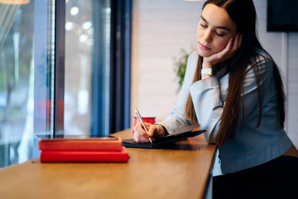 istock Thoughtful attractive student with pen in hand writing down poems in notebook sitting in coffee shop and enjoying tree time.Dreamy hipster girl noting text information in notepad and doing homework 908002462