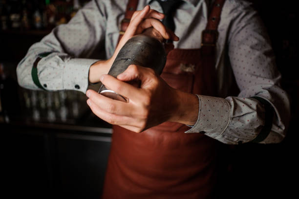 Barman in brown apron shaking the shaker Barman in a brown apron shaking the shaker on the dark background of bar counter cocktail shaker photos stock pictures, royalty-free photos & images