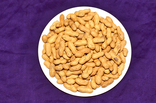 Roasted whole peanuts in white plate on a purple background.