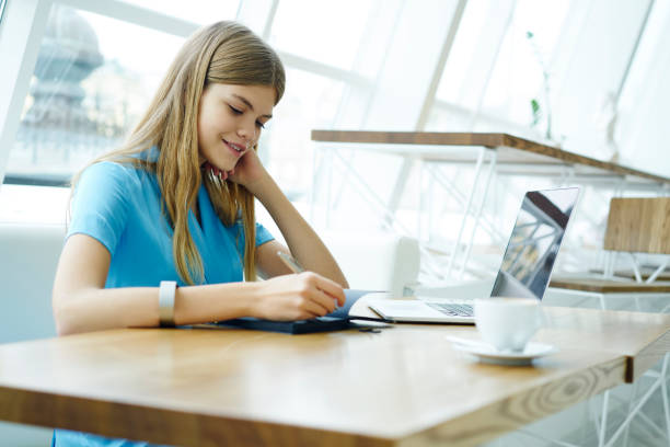 istock Smiling hipster girl recording notes in notebook sitting at laptop computer in university interior.Cheerful talented student writing down text information in notepad during preparation for exams 908006574