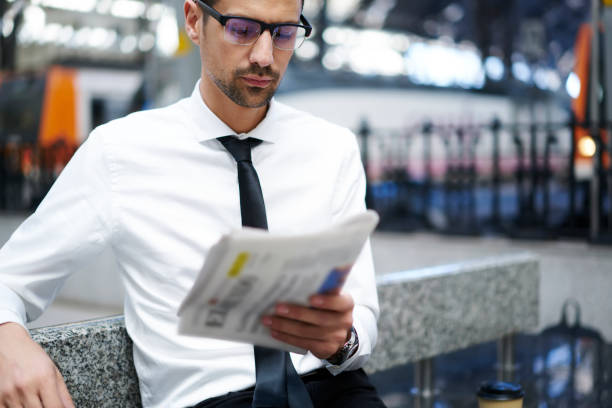 Pensive bearded manager in spectacles reading newspaper articles while waiting for public transport on subway, serious male in formal wear checking latest news from press sitting near copy space