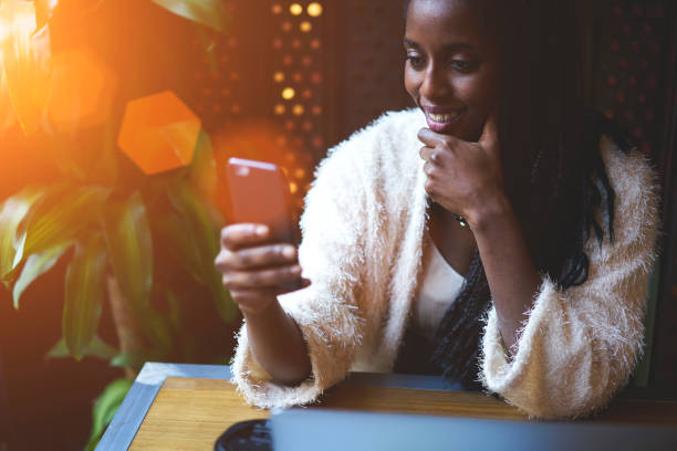 jeune noire modèle tenue trendy et bonne humeur te chat dans les réseaux sociaux à l’aide d’équipement moderne, adolescente, souriant tout en messagerie de rétroaction pour amis par le biais de smartphone assis au café à l’intérieur - lundi noir chinois photos et images de collection