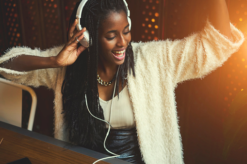 Young cheerful hipster female in casual wear listening music via big white modern headphones, afro american teen girl recreate while enjoying favorite songs from playlist sitting in cafe indoors