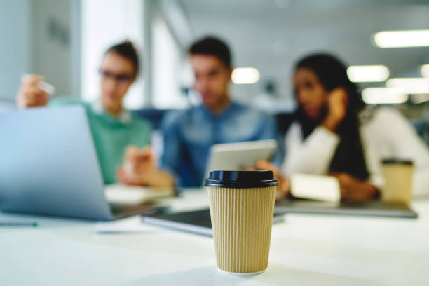 istock Selective focus on paper coffee cup standing on wooden table in coworking office during lunch break on blurred background with working people silhouettes sitting at desktop with modern technology 908020376