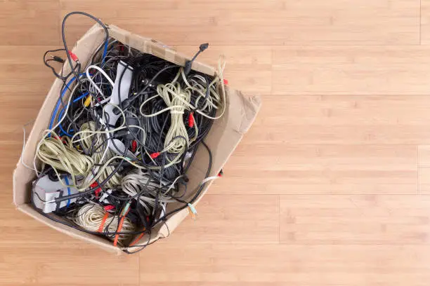 Old battered cardboard box with electrical cords and connectors for electronic devices on a wooden background with copy space viewed from above
