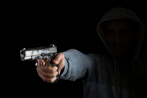 Gun in male human hand aiming toward left side in dark. The person is hidden in shadow, only hand and gun is illuminated. Shot indoor with a full frame DSLR camera.