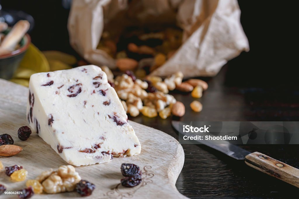 Wensleydale cheese with cranberries, red wine, honey, nuts, raisins on wooden cutting board. Black concrete background. Selective focus. Almond Stock Photo