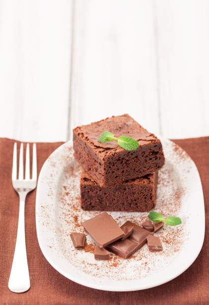 istock Chocolate brownie square pieces in stack on white plate decorated with mint leaves and cocoa powder on white vintage wooden background. American traditional delicious dessert. Close up photography 908044176