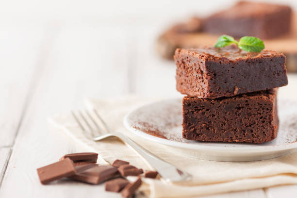 istock Chocolate brownie square pieces in stack on white plate decorated with mint leaves and cocoa powder on white vintage wooden background. American traditional delicious dessert. Close up photography 908044180