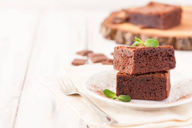 istock Chocolate brownie square pieces in stack on white plate decorated with mint leaves and cocoa powder on white vintage wooden background. American traditional delicious dessert. Close up photography 908044182