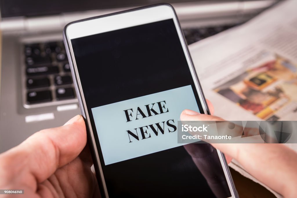 Man's hand holding a smartphone with Fake News words on screen. Fake news, HOAX concept Man's hand holding a smartphone with Fake News words on screen over a newspaper and a laptop. HOAX and Fake news concept. Fake News Stock Photo