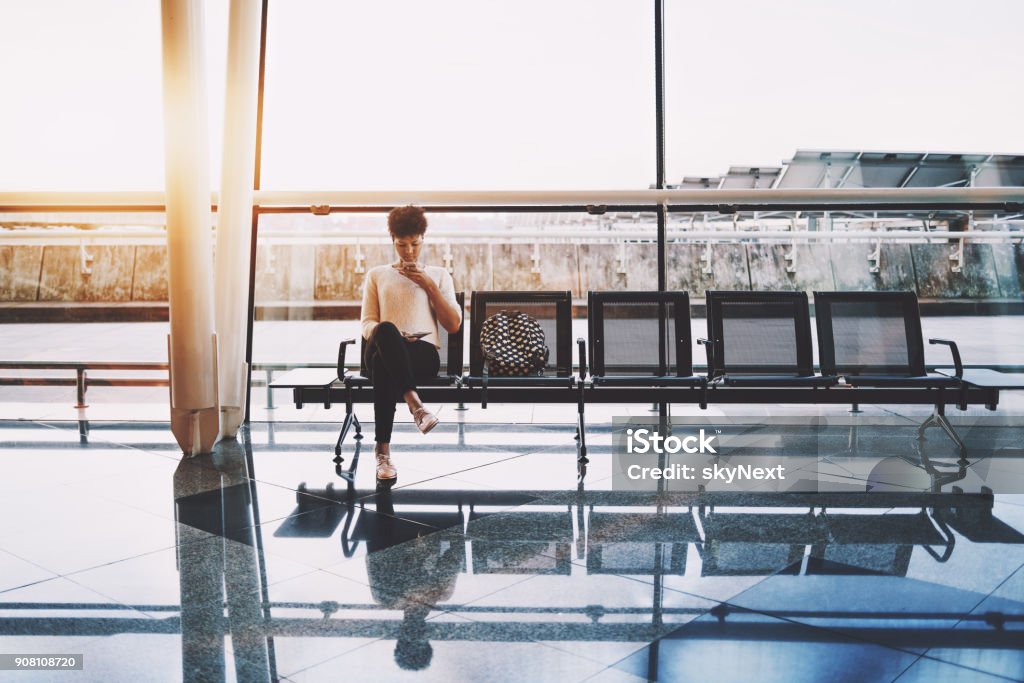 Black girl using cellphone in waiting hall of airport Cute Brazilian girl with curly Afro hair using smartphone for audio messaging while sitting on meatal seats of modern airport terminal or railway station, near huge window facade in background Airport Departure Area Stock Photo