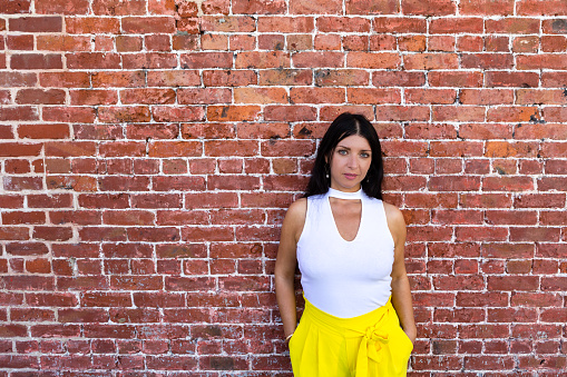 Serious fashionable young woman in yellow pants standing with her hands in her pockets in front of a brick wall with copy space