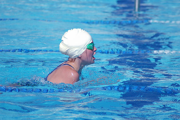 Young Female Swimmer swimming the Breaststroke Young teenage swimmer swimming the breaststroke. She is mid stroke with her head out of the water taking a breath. She is wearing a white swim cap and green rimmed swim goggles. She is 12-13 years old. swimming pool lane ropes stock pictures, royalty-free photos & images