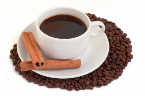 A cup of black coffee with two cinnamon sticks, surrounded by roasted coffee beans.