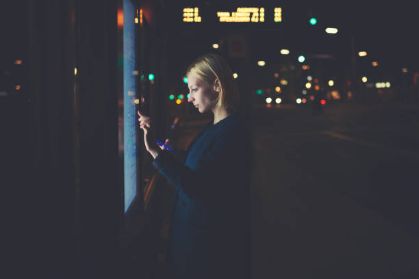 istock Young blonde female using tourist information self service while standing in beautiful night city with lights on background, pretty woman touching big digital screen of smart bus station of Barcelona 908210318