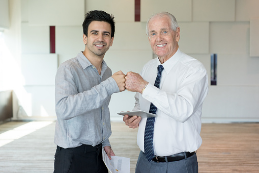 Two smiling business partners bumping fists in winning gesture. Senior businessman holding tablet and greeting his partner. Business meeting and success concept