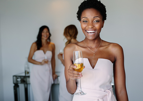 Beautiful young bridesmaid having wine on wedding day. Smiling african woman having drinks in a hotel room.