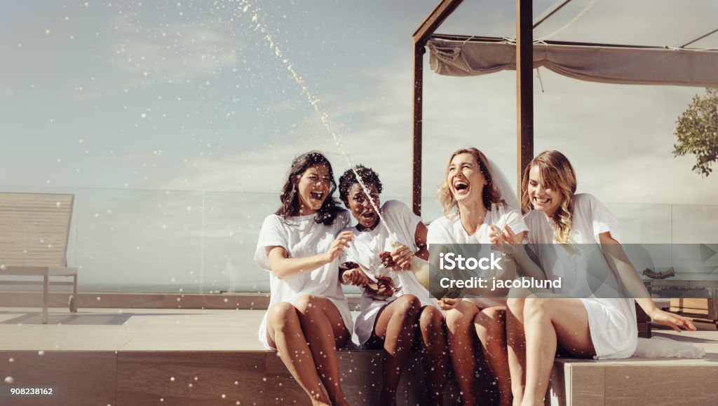 Cheerful bride and bridesmaids celebrating hen party Cheerful bride and bridesmaids celebrating hen party with champagne while sitting on rooftop. Girls having a great time at the hen party. Bachelorette Party Stock Photo