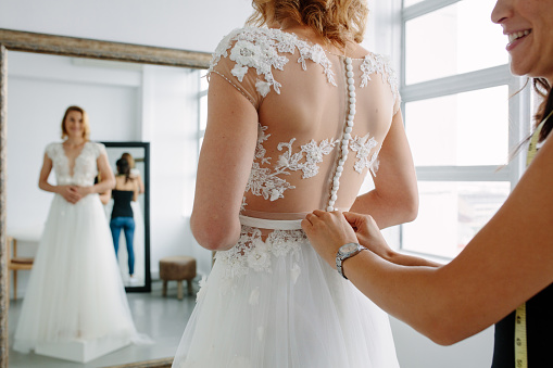 Bride is standing in front of mirror and fitting dress in atelier with wedding assistant. Woman with dressmaker are making final touch on tailor made gown in bridal clothing shop.