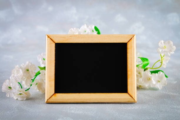 istock Chalkboard in a wooden frame with an empty box surrounded by white flowers on a gray background. Copy the space. Template for spring or female holiday. 908244304