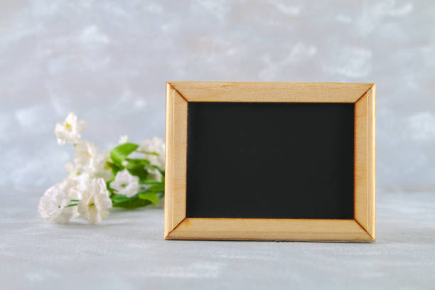 istock Chalkboard in a wooden frame with an empty box surrounded by white flowers on a gray background. Copy the space. Template for spring or female holiday. 908244316