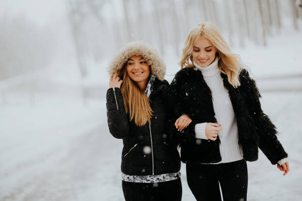 Three long-haired young women dressed in a casual outerwear are standing together outdoors on snow-covered playground. They are looking at the camera. Shooting at winter day in a courtyard of a residential building