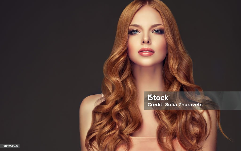 Young, red haired beautiful model with long, curly, well groomed hair. Excellent hair waves. Young, red haired woman  with curly and voluminous hair. Beautiful model with long, dense wavy hairstyle and vivid make-up. Perfect hair waves and passionate look. Hairstyle Stock Photo