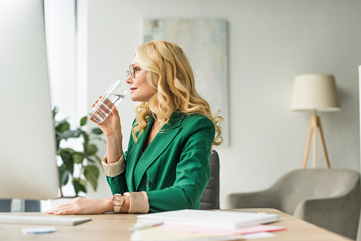 mature businesswoman in eyeglasses drinking water from glass and looking away