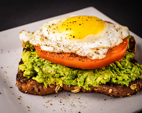 Avocado toast on white plate made with whole grain bread, sunny side up fired egg, and a sliced tomato.