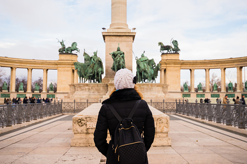 Young woman is standing in Heroes square and observing the monument in BudaPest, Hungary. The sculptures were made by sculptor Zala György from Lendava in 1896 in Budapest.
