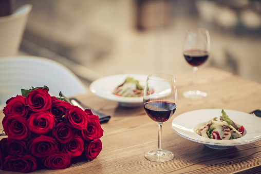 Romantic dinner settings. Two glasses of wine, dishes and bouquet of red roses are on table.