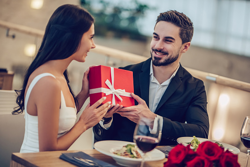 Beautiful loving couple is spending time together in modern restaurant. Attractive young woman in dress and handsome man in suit are having romantic dinner. Celebrating Saint Valentine's Day.