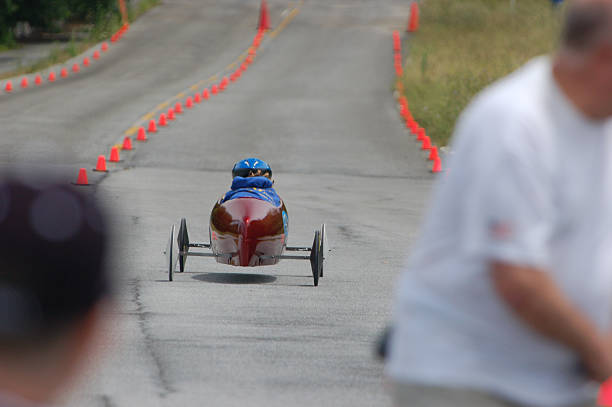 Race On Youth in a soap box race car speeds down the track. Photo taken from just behind the starting line. DOF focus on race car driver while out of focus race officials frame the subject left and right. soapbox cart stock pictures, royalty-free photos & images