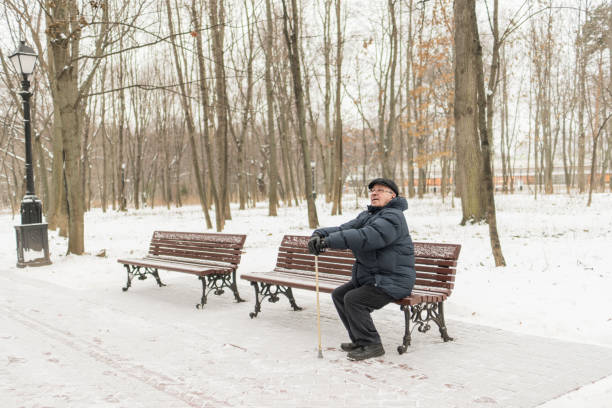 istock Man - elderly pensioner, sits on a park bench in the winter. High trees, a lantern. 908352728