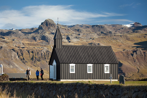 Located on Iceland’s Snaefellsnes Peninsula near the fishing village of Budir, visitors explore the black Church of Budir which stands with snow capped mountains in the distance and a walled graveyard in the foreground.