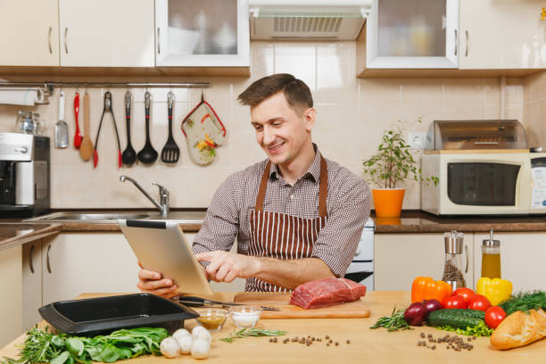 istock Young man in apron sitting at table with vegetables, looking for recipe in tablet, cooking at home preparing meat stake from beef, in light kitchen with wooden surface, full of fancy kitchenware. 908389362