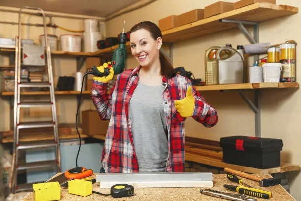 Beautiful caucasian young brown-hair woman in plaid shirt and gray T-shirt working in carpentry workshop at table place, drilling with drill holes in piece of iron and wood while making furniture